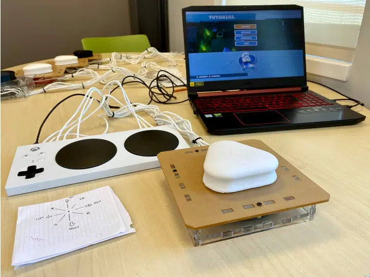 A table with an open laptop in the center wired to an Xbox Adaptive Controller and the controller developed by UPEI students wired to the Xbox Adaptive Controller